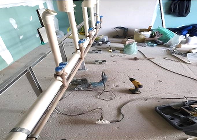 Tubulações de PVC com registros azuis montadas sobre suporte metálico, em obra com ferramentas e cabos no piso